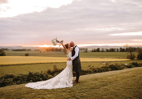 a bride wearing a lace dress and groom wearing a kilt kiss in a field of grass at sunset. The bride is holding her bouquet in the air