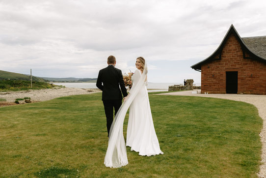 A bride and groom stroll hand in hand on lush grass towards the sea with a red brick building with pitched roof on right