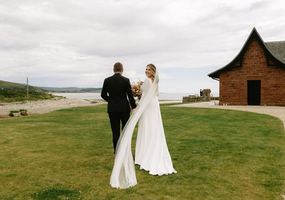 A bride and groom stroll hand in hand on lush grass towards the sea with a red brick building with pitched roof on right