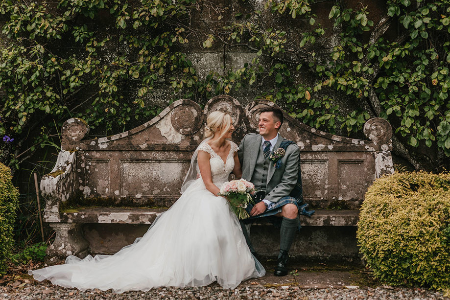 A bride in a white dress and a groom in a grey kilt sit on a stone bench 