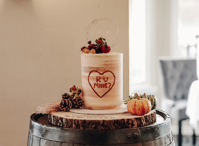 a marbled tall one-tier wedding cake decorated with 'RU MINE' lettering within a heart. It sits on top of a rustic wooden barrel surrounded by pine cones and mini pumpkins