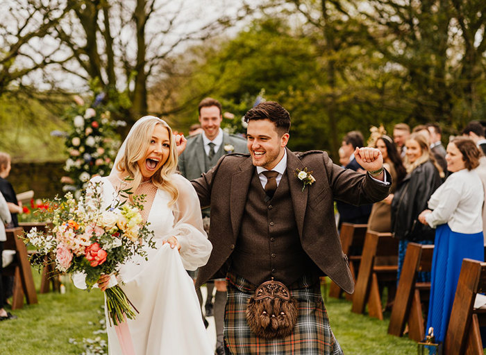 an elated newlywed bride and groom cheer in a garden with trees and flowers in background. Wedding guests are seated on rows of wooden folding chairs either side of the couple