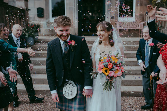 a bride and groom walking through a colourful confetti shower outside Netherbyres House