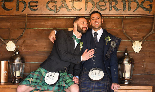 Two grooms wearing Highlandwear sitting on wooden bench with wooden sign saying 'The Gathering'