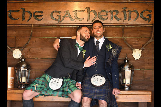 Two grooms wearing Highlandwear sitting on wooden bench with wooden sign saying 'The Gathering'