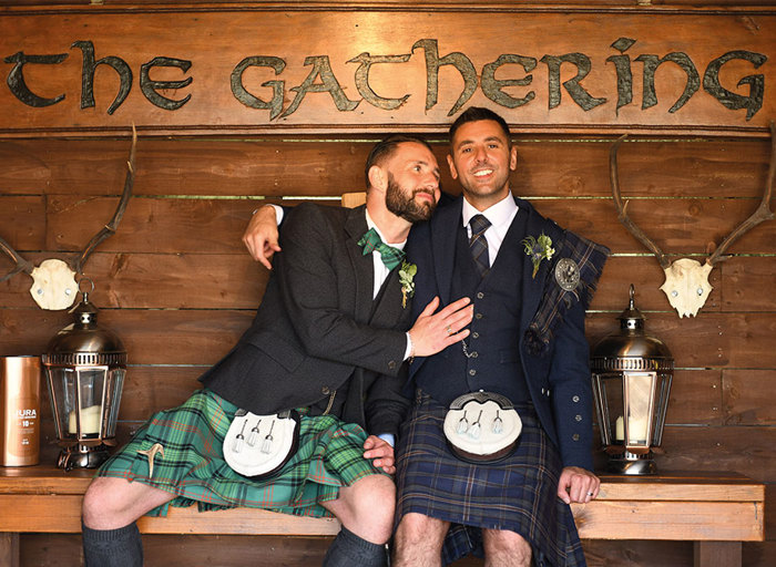 Two grooms wearing Highlandwear sitting on wooden bench with wooden sign saying 'The Gathering'