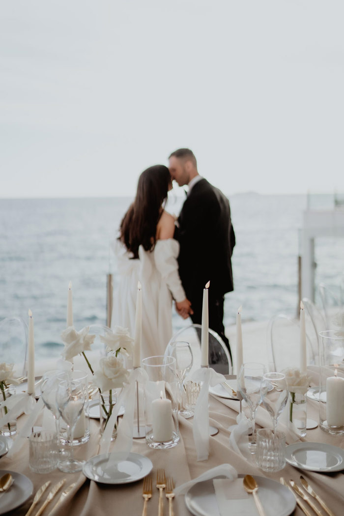 a couple kissing with the sea in the background. An elegant table set for a wedding with tall slim white candles, white roses, gold cutlery and a peach table cloth