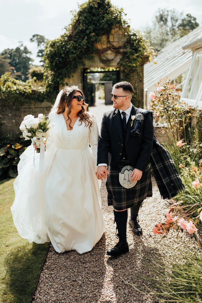 A man in a dark kilt holding hands with a bride in a long sleeved wedding dress as they  walk along a path through a garden