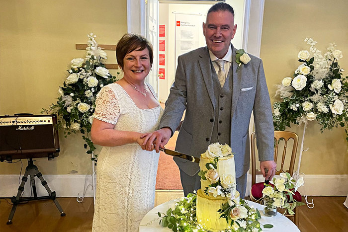 A bride and groom cut their two tier wedding cake which is covered in greenery and white flowers 