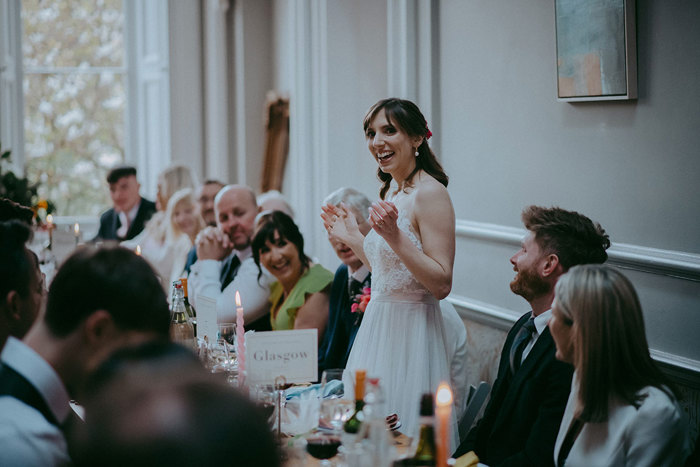 a bride standing making a wedding speech at a long table as other guests watch on