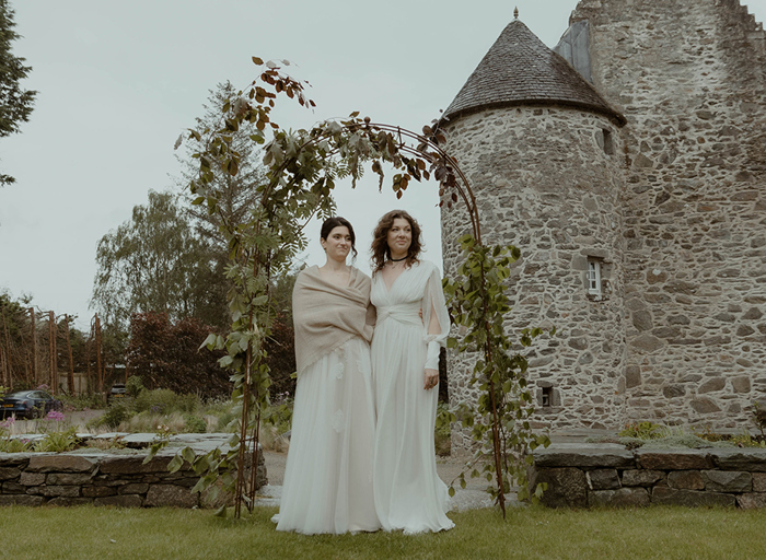 two brides standing outside under a foliage-covered wire arch in a garden with old stone building in background