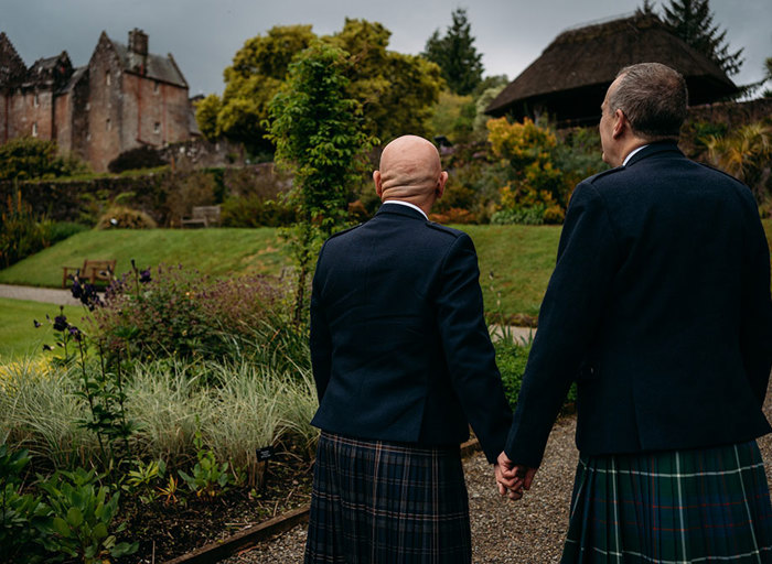 two grooms wearing kilts hold hands in a garden setting and look towards Brodick Castle