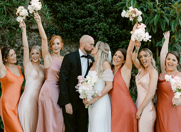 a bride and groom kiss in front of a green hedge amid a lineup of bridesmaids wearing dresses in shades of orange, terracotta, pink and oyster