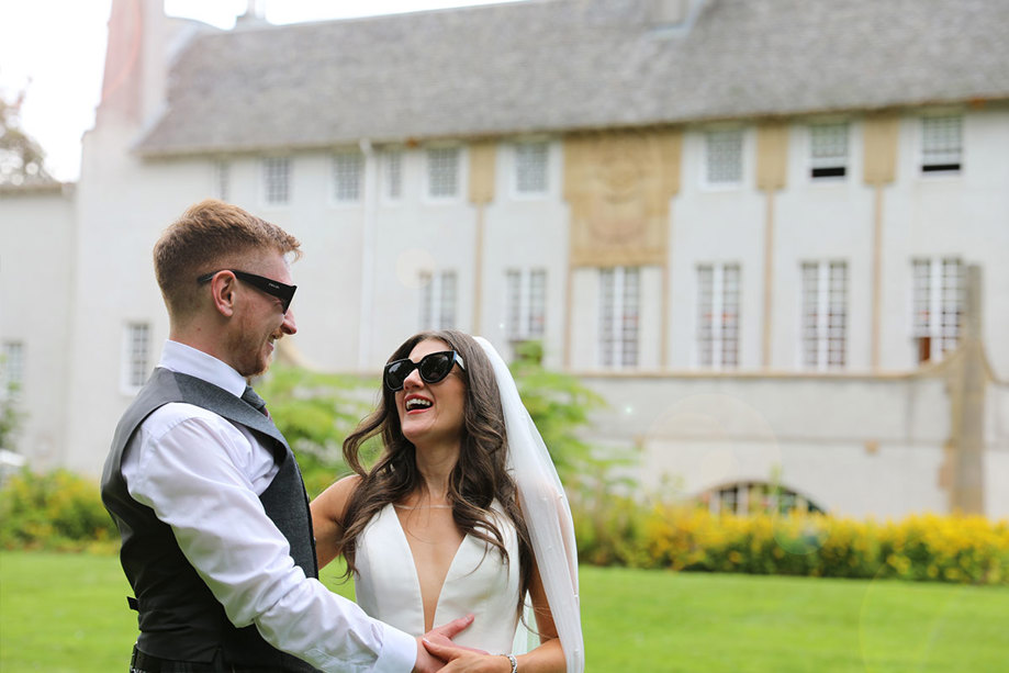 A bride and groom laughing while looking at each other and wearing sunglasses 