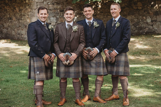 Four men standing outside wearing matching brown kilts, three with navy jackets and one with a brown jacket