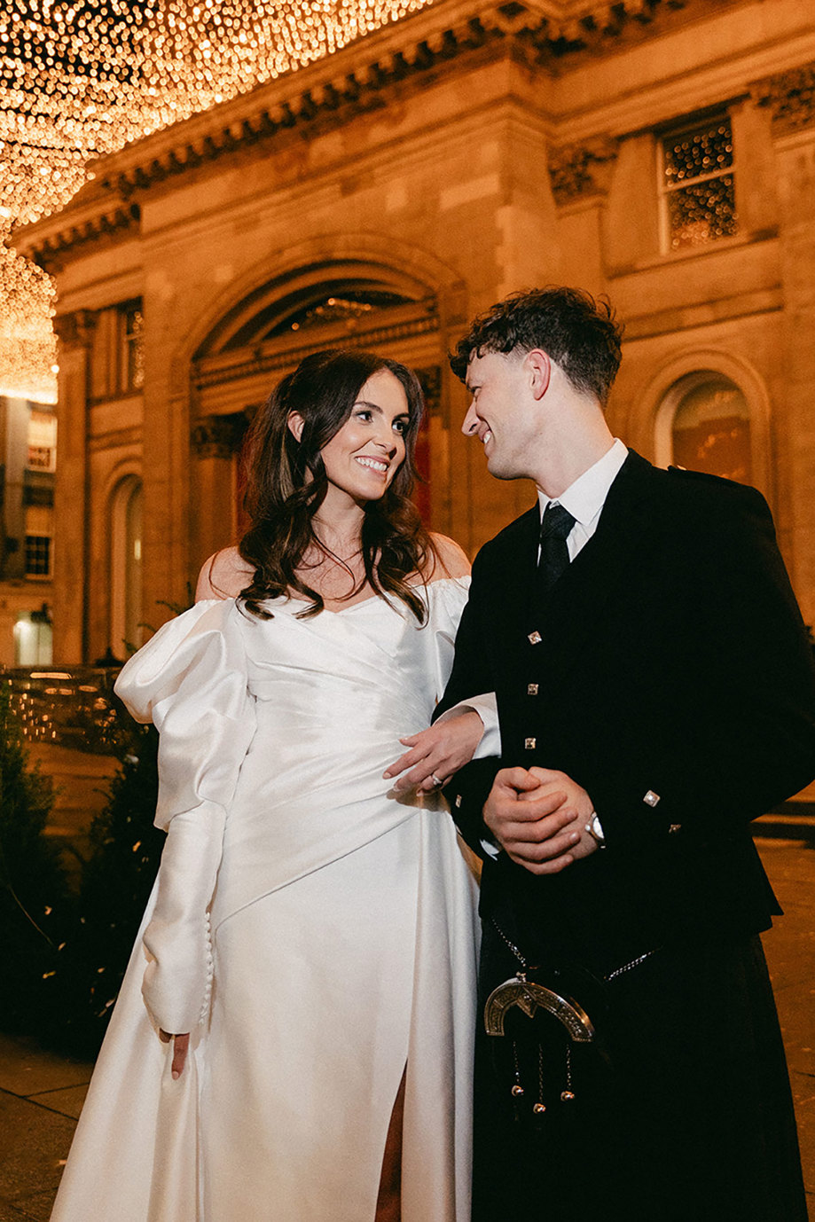 brunette bride in high leg slit wedding dress with long puff sleeves and groom in full dark tartan kilt outfit smile at each other outdoors with a canopy of twinkling lights in the night sky above them