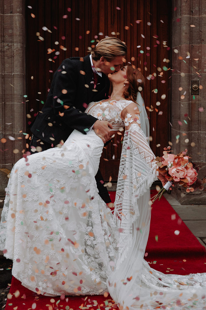 groom dips bride in in flowy bohemian wedding dress for a kiss as they are surrounded by colourful confetti and stood on a red runner