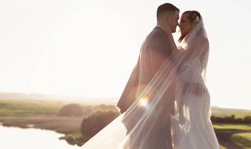 a bride and groom standing nose to nose basking in a golden sunset with green landscape and lake in background