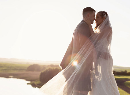 a bride and groom standing nose to nose basking in a golden sunset with green landscape and lake in background