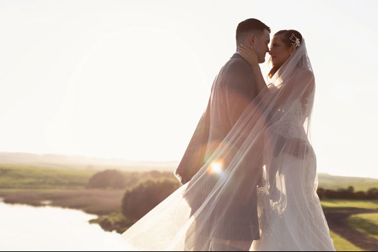 a bride and groom standing nose to nose basking in a golden sunset with green landscape and lake in background