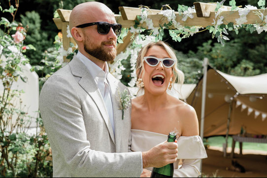 an elated bride and groom pop a champagne bottle in a garden setting with canvas stretch tent, wooden structure and greenery in background