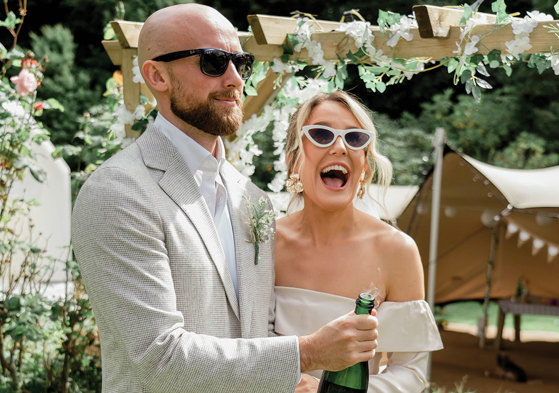 an elated bride and groom pop a champagne bottle in a garden setting with canvas stretch tent, wooden structure and greenery in background