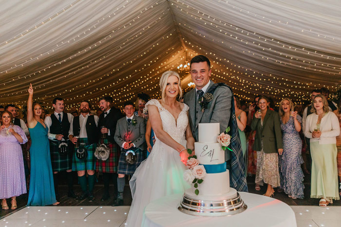 A bride and groom cut their three tier white wedding cake which has pink roses and a blue ribbon 