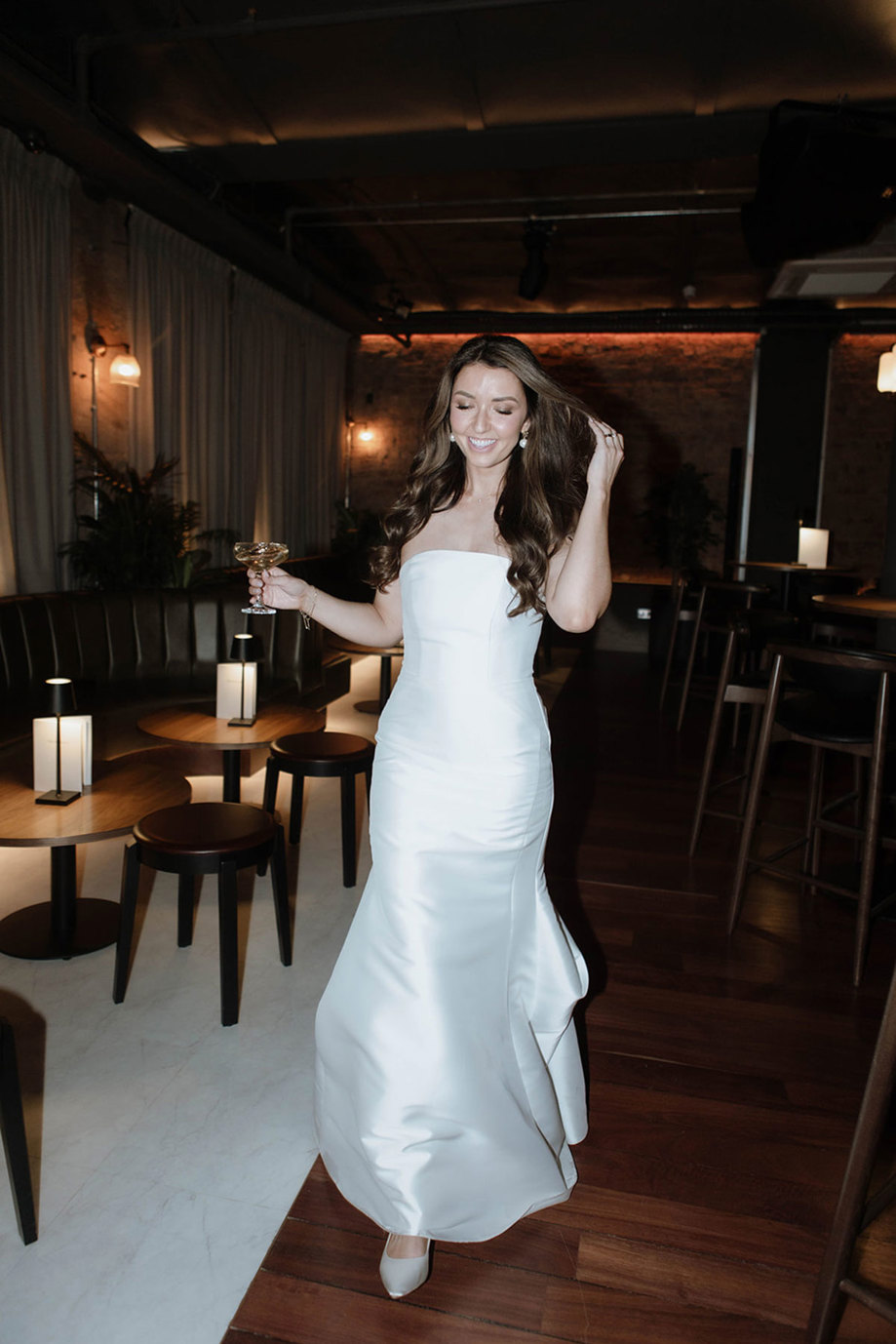 A bride wearing a fitted satin gown walks towards the camera holding a champagne coupe