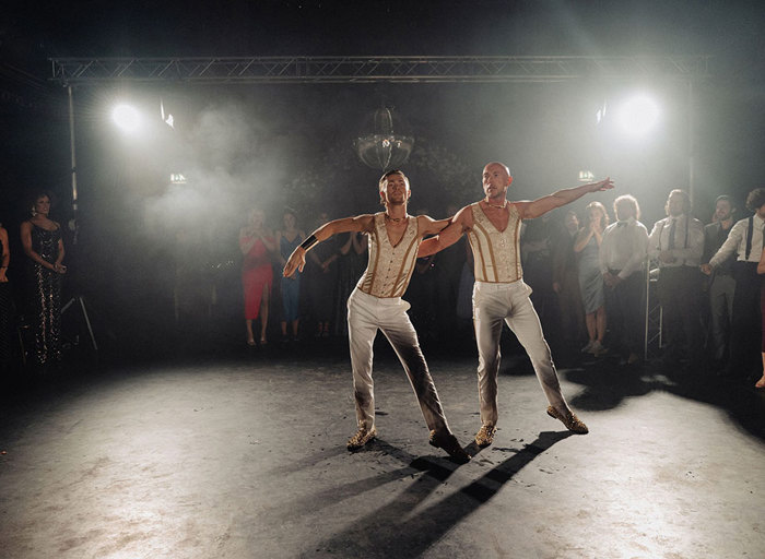 Grooms in matching cream and white trousers dancing surrounded by crowd 