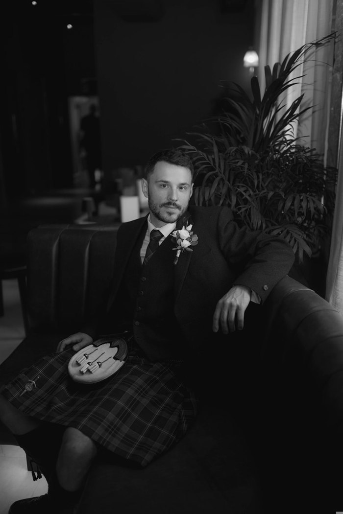 A black and white photo of a man wearing a kilt sitting on a couch and looking at the camera