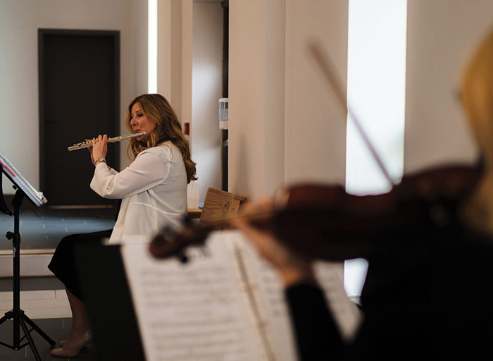 a person playing a flute reading sheet music from a music stand with a blurred out person playing a violin in foreground