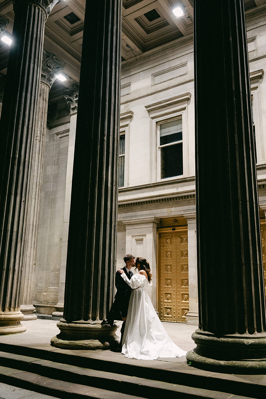 brunette bride in trailing wedding dress with long puff sleeves and groom in full dark tartan kilt outfit kiss outdoors stood between two large historic columns