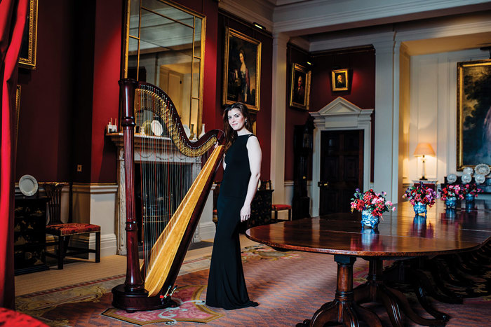 a brunette woman in a long black gown stands side by side with a large harp within a traditional stately dining room