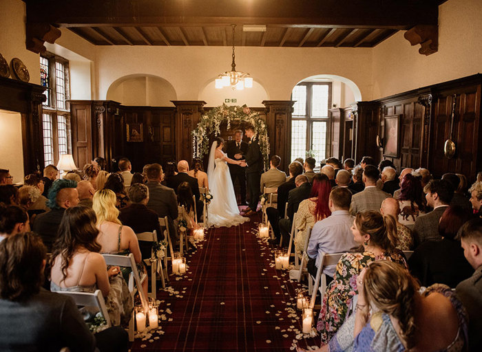 A wedding ceremony in a dark wooden panelled room looking up the aisle at the bride and groom holding hands