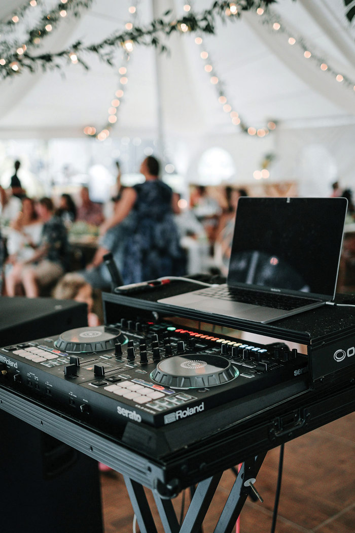 a DJ turntable and laptop are perched at the front of photo and blurred people and fairylights strung from the top of marquee are visible in the background