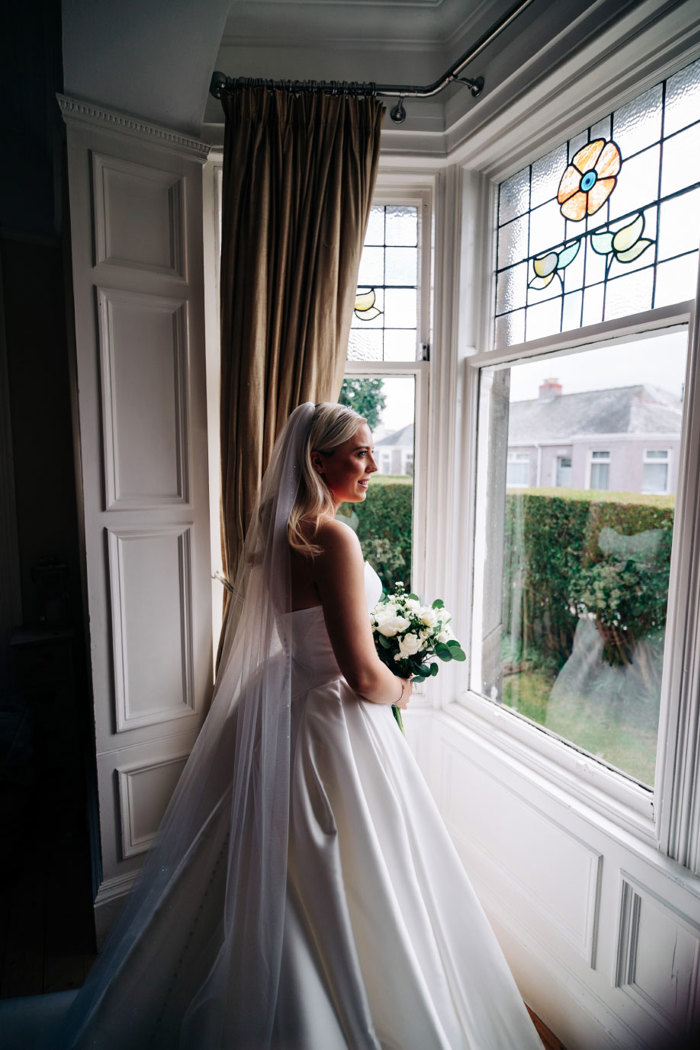 a bride wearing a veil and ivory strapless dress and holding a bouquet while standing by a window looking out onto a hedge and green garden