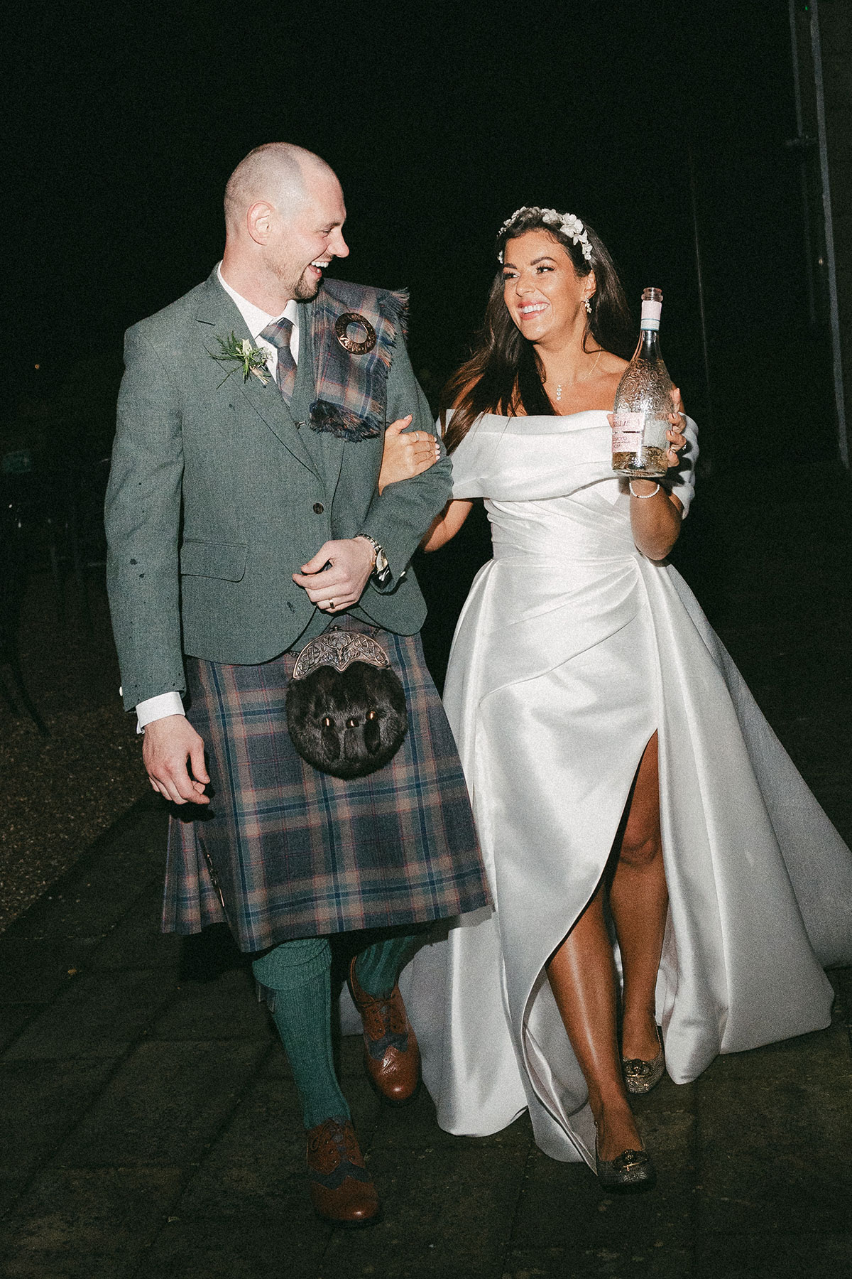 bride with floral headband and off the shoulder wedding dress with a leg slit holds prosecco bottle in one hand and groom in green, blue and purple tartan kilt outfit with green jacket in the other