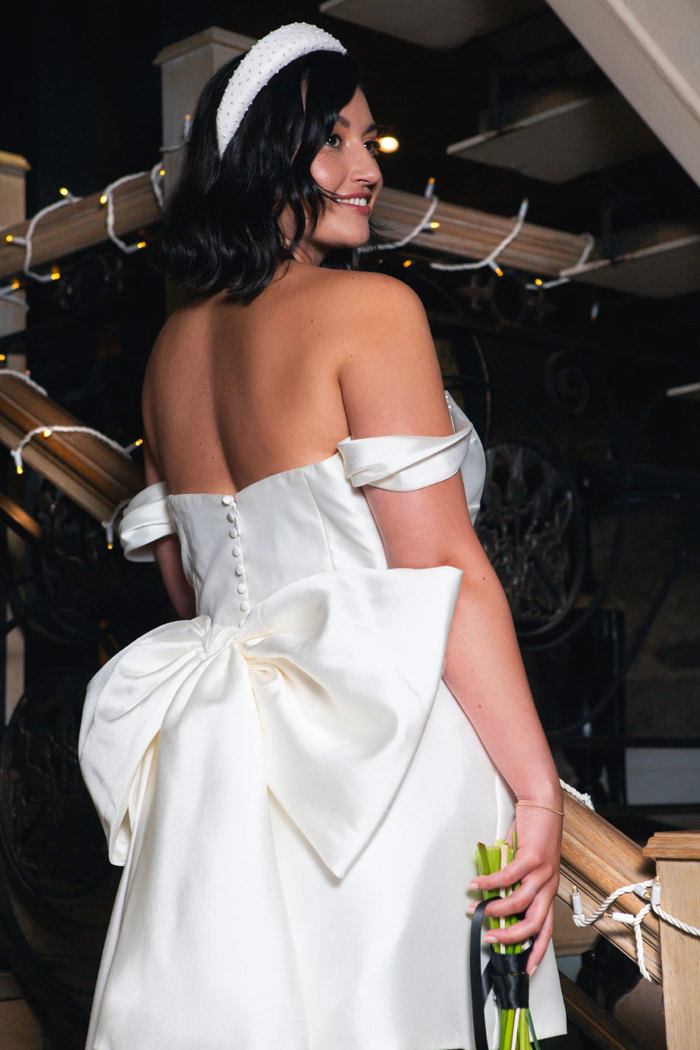 A woman facing away from the camera and looking over her shoulder wearing a white satin dress with a large bow in the back and a beaded white headband 