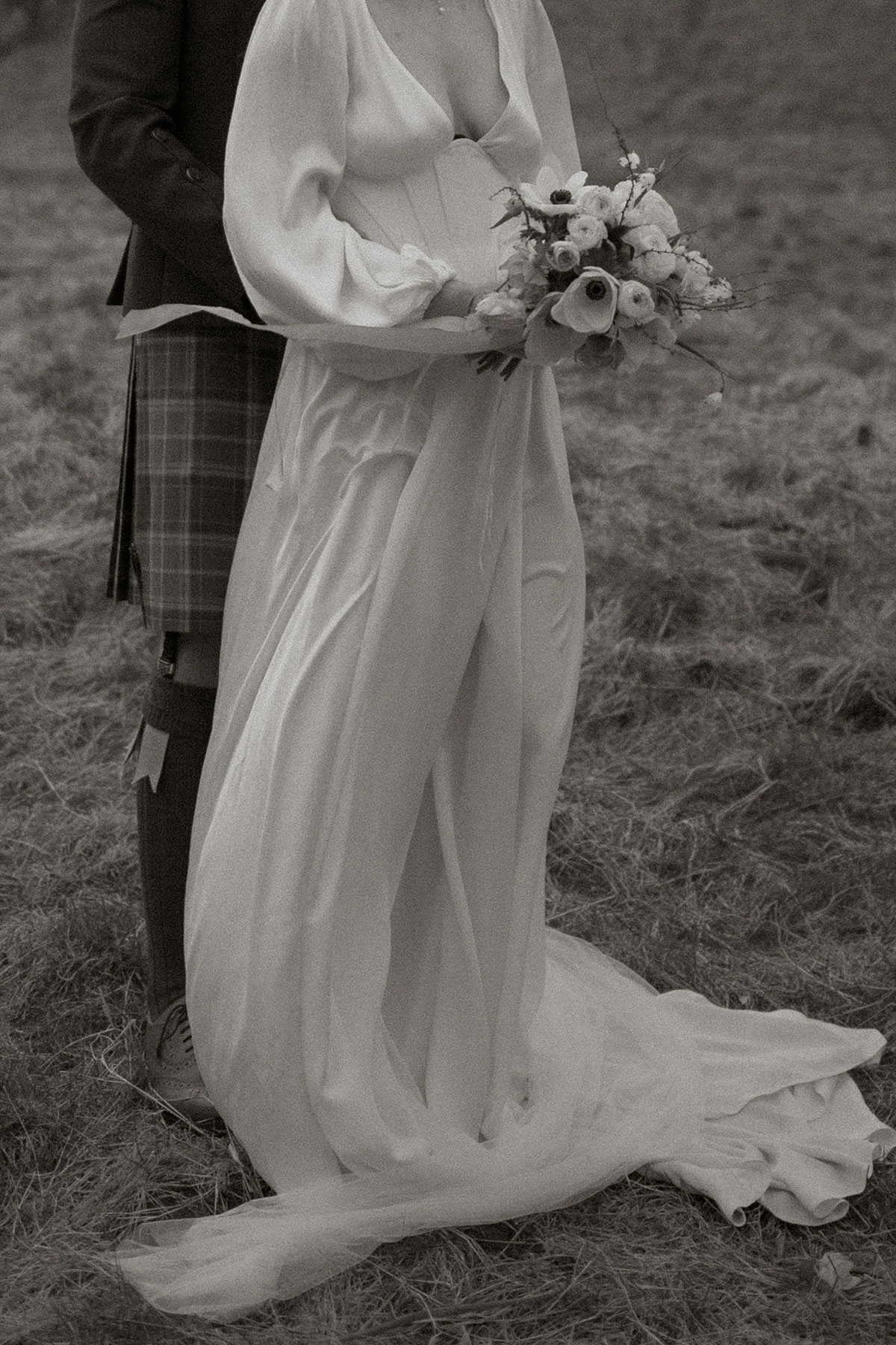 black and white photo of bride in plunging satin dress holding a bouquet of flowers, standing in front of groom in kilt outfit, both captured from the neck down, stood outdoors on the grass