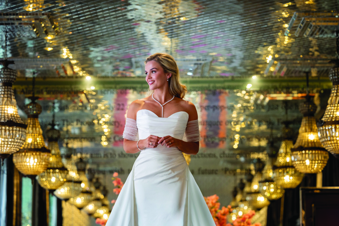 A woman wearing a strapless wedding dress with tulle sleeves standing in a mirrored room with crystal chandeliers