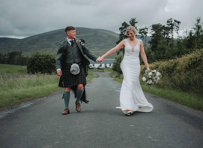 bride and groom walking hand in hand along a countryside road with hills and greenery in background