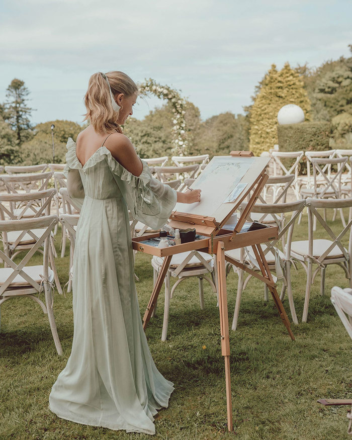 a blonde woman in a floaty green dress stands at an easel, painting a wedding ceremony set up