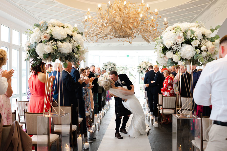 a groom dips a bride to kiss her while rows of guests cheer and clap. They are standing below an ornate chandelier and in front of them lies tall round floral arrangements on perspex plinths.