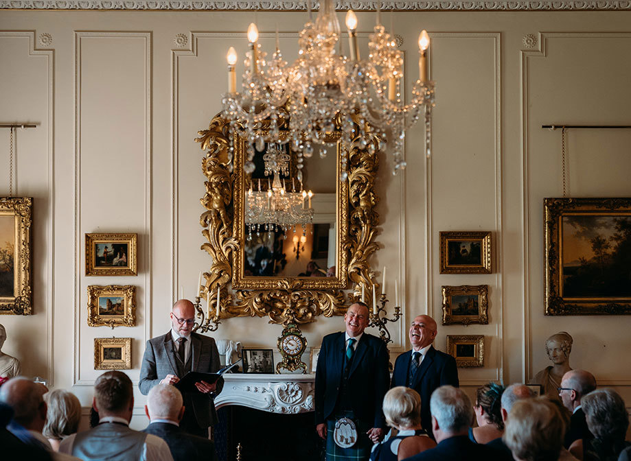 a wedding ceremony of two laughing grooms in a grand room with large gold framed mirror and chandelier at Brodick Castle