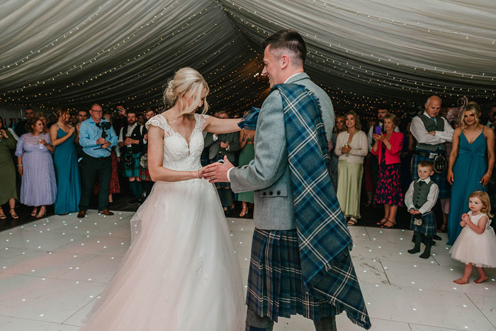 A bride and groom dance together on a white dance floor with their guests gathered around them and fairylights above them