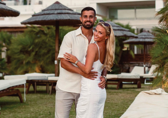 a smiling man and woman posing for a photograph in front of sunbeds and sun parasols with greenery in the background