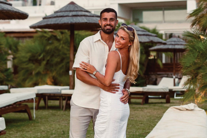 a smiling man and woman posing for a photograph in front of sunbeds and sun parasols with greenery in the background