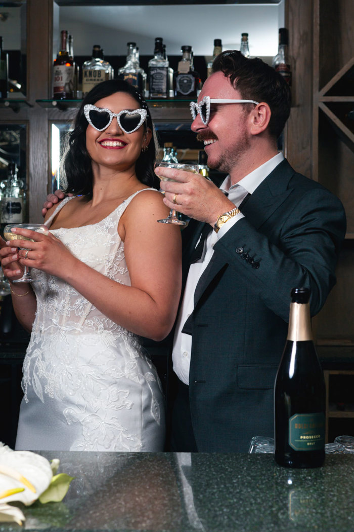 A woman in a white sparkly dress and a man in a suit with his bowtie undone hold up champagne coupes while wearing white heart shaped sunglasses