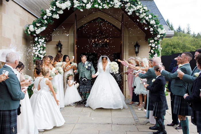 a bride and groom emerging from a doorway. Two rows of guests throw white confetti towards them.