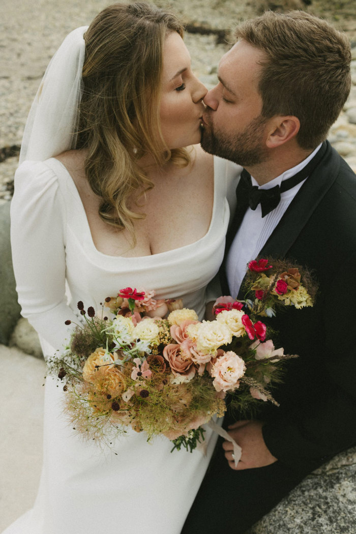 a bride holding a bouquet of flowers and wearing a square-neck white dress kissing a groom wearing a smart black tie suit